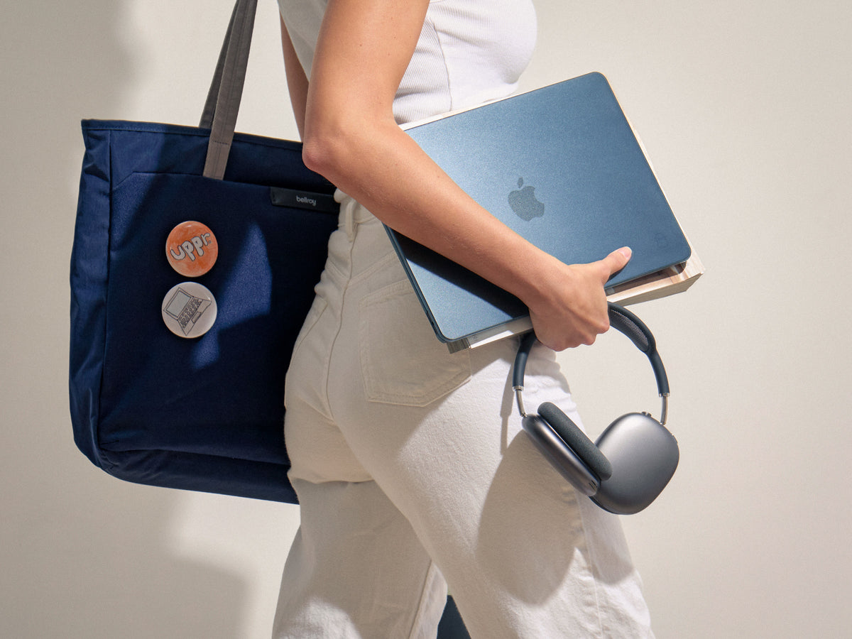 Person carrying a Midnight MacBook Air under one arm with a navy tote bag and headphones